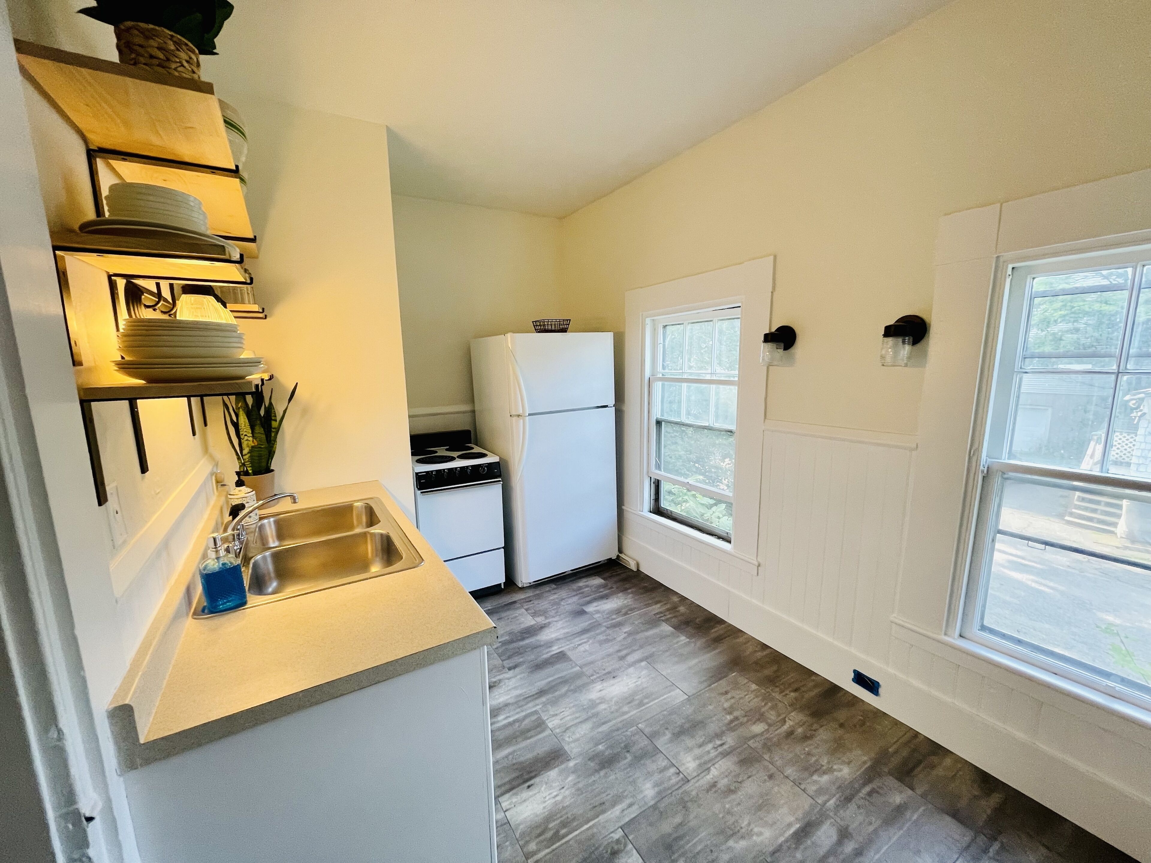 A compact kitchen with a sink, stove, and refrigerator, wooden shelves, and a window providing natural light.