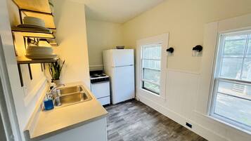 A compact kitchen with a sink, stove, and refrigerator, wooden shelves, and a window providing natural light.