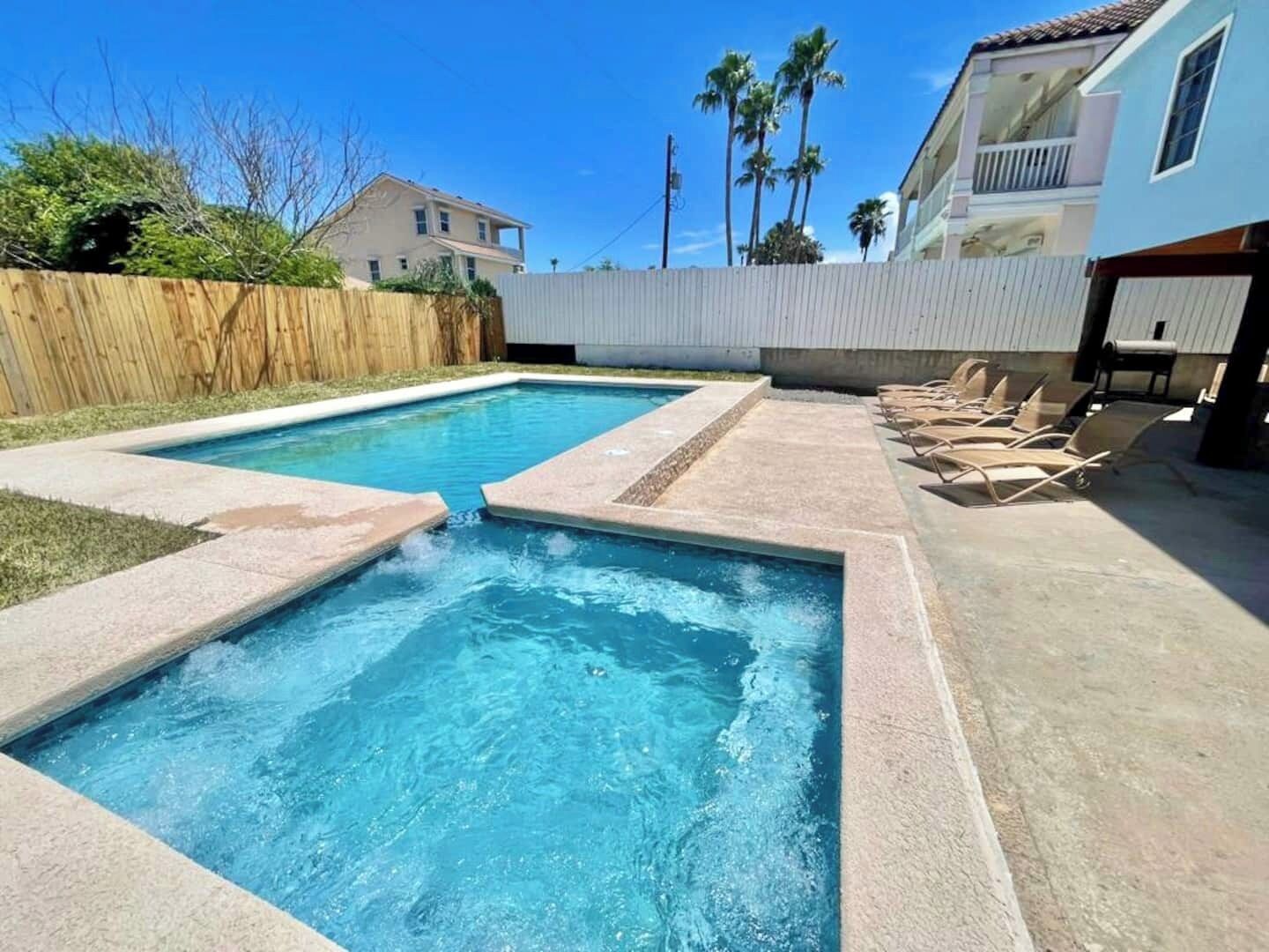 Una piscina rectangular con una pequeña cascada, rodeada de una terraza de hormigón y sillones de descanso.