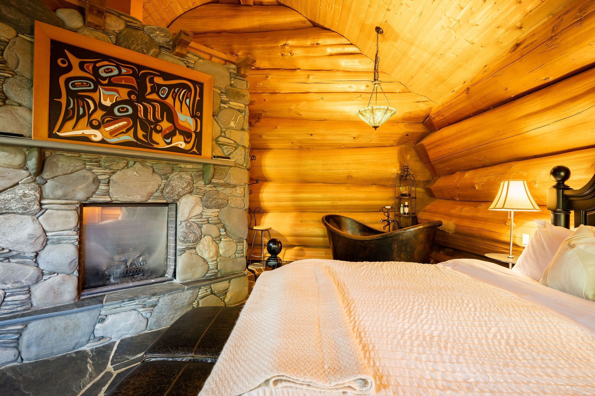 A log cabin room with a stone fireplace, a bathtub, and a bed with a white blanket.