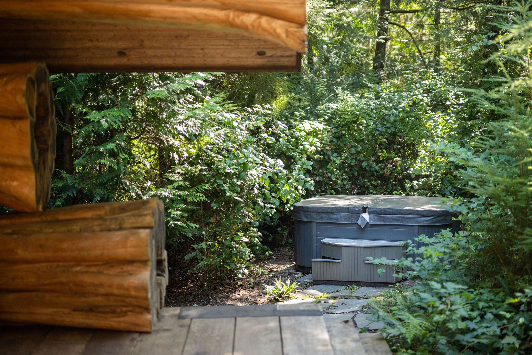 Un bain à remous entouré d’une végétation luxuriante, avec des rondins de bois et un sentier en pierre.