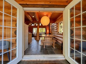 L’intérieur d’une cabane en rondins, avec une table et des bancs en bois au centre, une lanterne suspendue et une vue sur la forêt environnante.