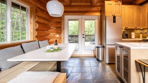 A modern log cabin with a large dining table, built-in bench seating, a kitchen with wooden cabinets, and a view of the surrounding forest through the glass doors. - Willowbrae Chalet-Sophisticated Forest-Hot Tub (Ucluelet)