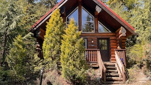 A log cabin with a red roof, large windows, and a wooden staircase leading to the entrance. - Willowbrae Chalet-Sophisticated Forest-Hot Tub (Ucluelet)