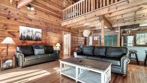A wooden cabin interior with two leather sofas, a white coffee table, and a kitchen area.