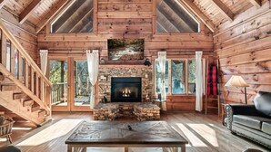 A log cabin interior with a stone fireplace, wooden floors, and a large window overlooking a deck.