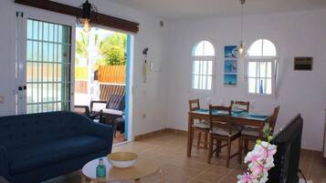 A living room with a sofa, a dining table with chairs, and a TV. There is a sliding glass door leading to an outdoor area.