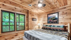 Een houten slaapkamer met een bed, een raam met zonwering, een plafondventilator, een wandlamp en een lijst met een afbeelding.