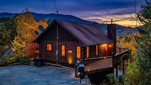 Een houten chalet met een veranda, omringd door herfstkleurige bladeren en heuvels.
