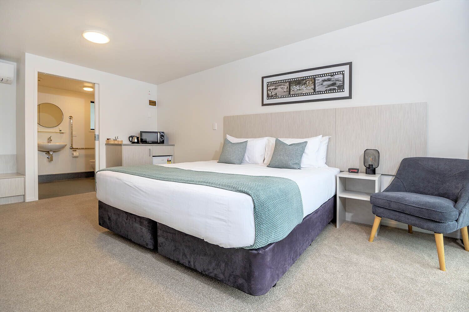 A modern hotel room with a large bed, a bedside table, a chair, a microwave, and a framed picture on the wall.
