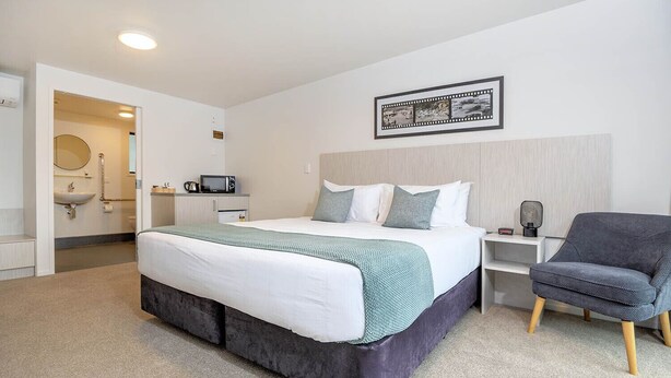 A modern hotel room with a large bed, a bedside table, a chair, a microwave, and a framed picture on the wall. - The Com Plex Motel (Whakatane)