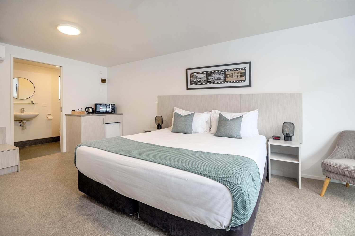 Ein modernes Hotelzimmer mit einem großen Bett, einem Nachttisch, einem Sessel, einem Schreibtisch und einem durch eine geöffnete Tür einsehbarer Badezimmer.
