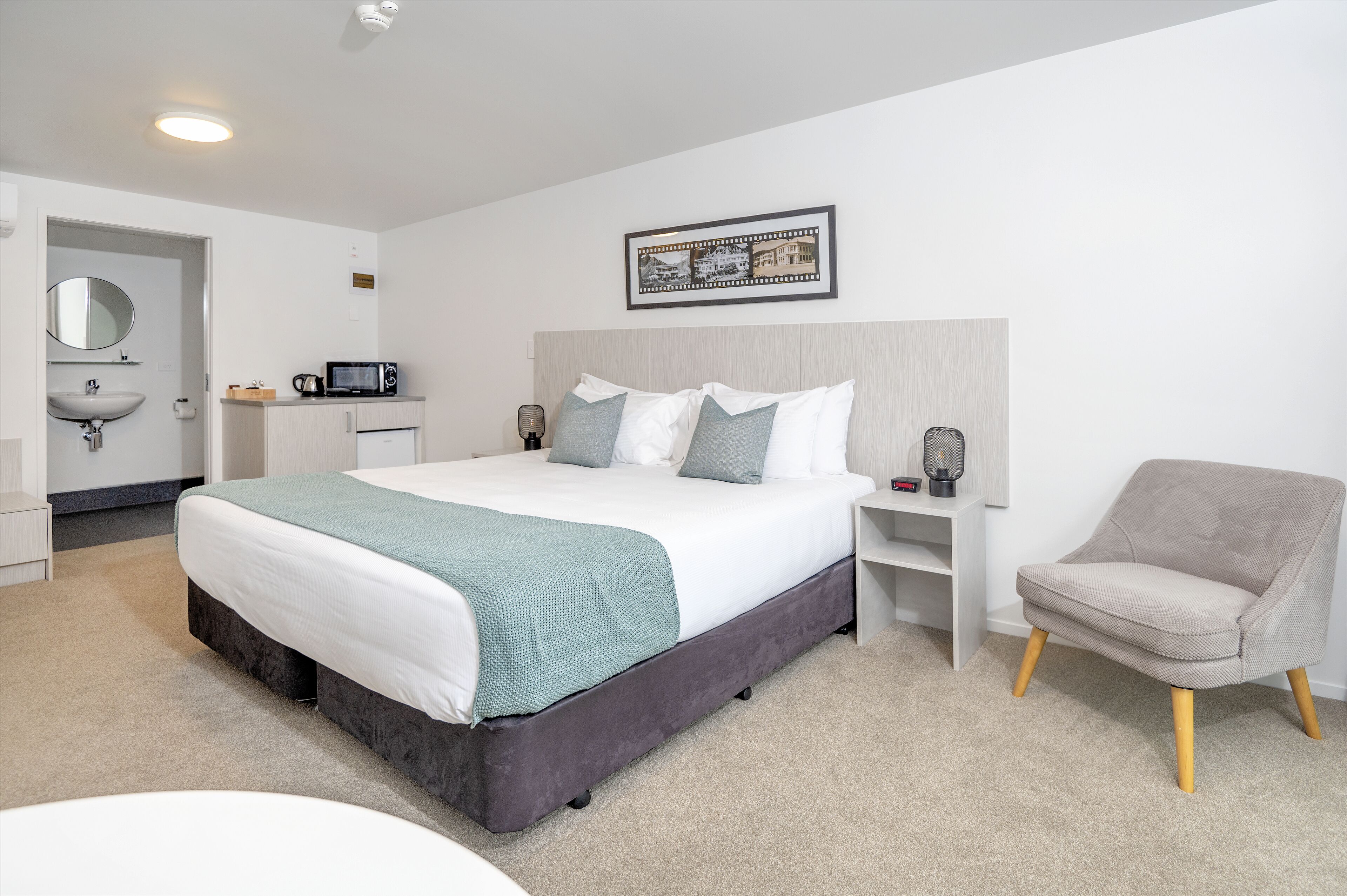 A modern hotel room with a large bed, bedside tables, a chair, a desk, and a bathroom with a sink and mirror.