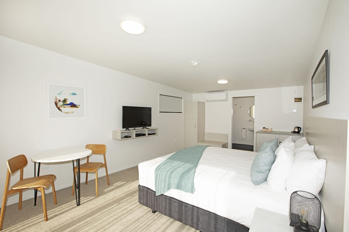 Habitación de hotel con cama, escritorio, dos sillas, televisor y un baño visible a través de una puerta abierta.