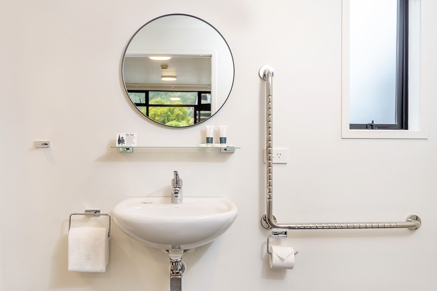 A bathroom with a white sink, a round mirror, a towel rack, a grab bar, and a small shelf with toiletries.