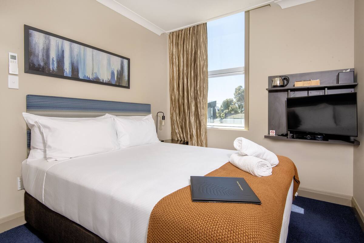 Una habitación de hotel con una cama, un cuadro en la pared, un televisor y una ventana con cortinas.