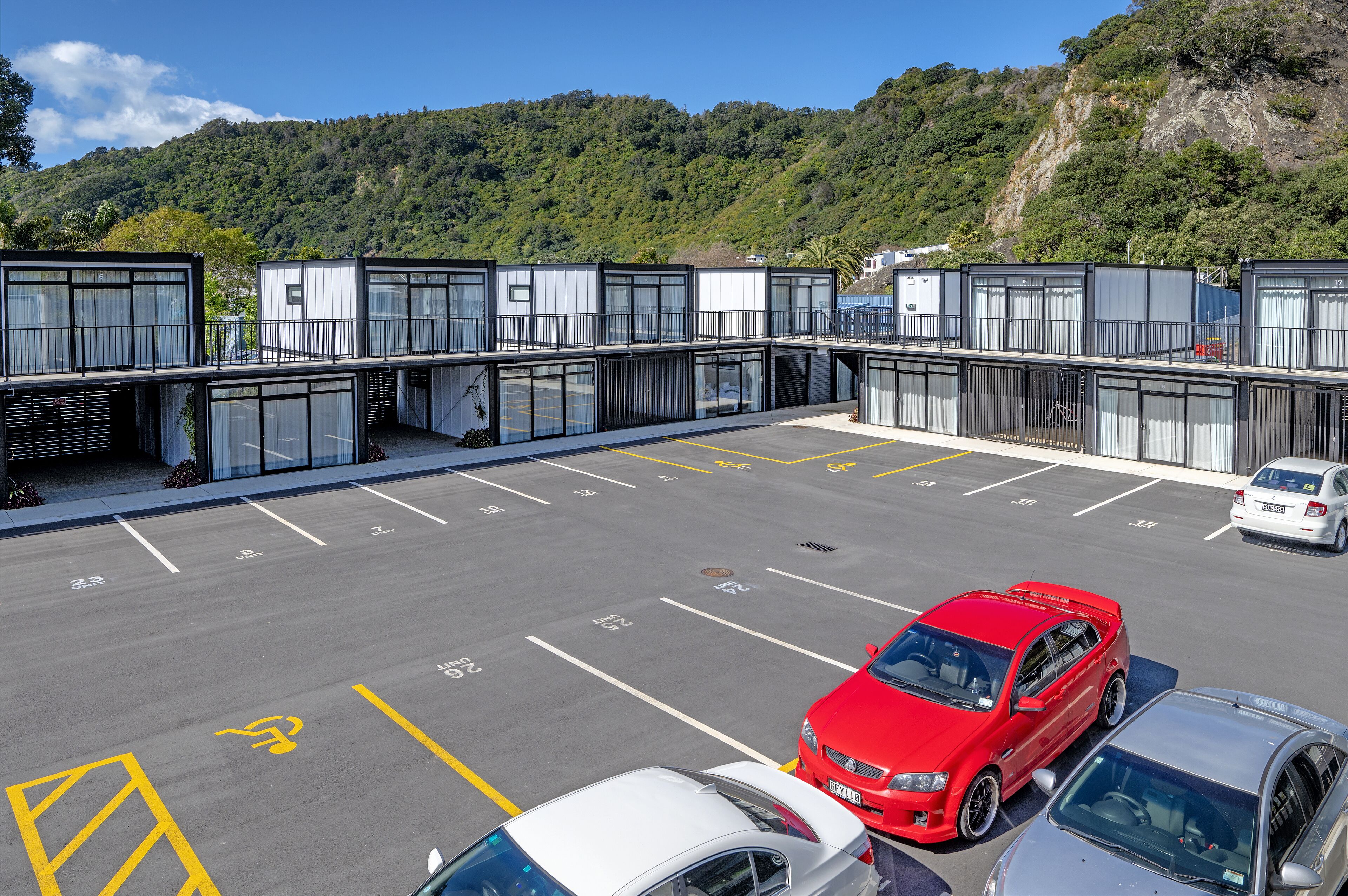 Um estacionamento moderno com vários carros, incluindo um carro esportivo vermelho, e um edifício com janelas amplas e sacadas.