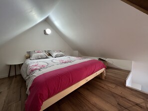 A bedroom with a sloped ceiling, a wooden bed frame, a small bedside table, and a wall-mounted light fixture. - Lapolline (Wimereux)