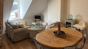 Camera da loft accogliente con soffitto spiovente, una grande finestra, un tavolo da pranzo in legno, un divano, una TV e una seggiolina per neonati.
