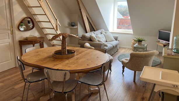 Una stanza in stile loft con un tavolo da pranzo in legno, sedie, un divano e un televisore. Si intravede una scala in legno e un lucernario.
