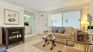 A living room with a sofa, coffee table, and a bookshelf. There is a kitchen area visible through an open door. - A Gem of a Studio at The Diamond Head Beach Hotel (Honolulu)