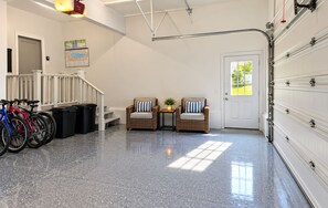 A clean, well-lit garage with a white door, a set of stairs leading to a second floor, and two wicker chairs with cushions.