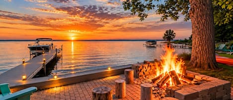 A waterfront deck with a fire pit, docked boat, and a view of the sunset over water.