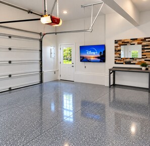 A spacious garage with a polished floor, a closed garage door, a mounted television displaying a Disney+ channel, and a wall-mounted shelf with a plant. - Waterfront Oneida Lake Gem House (Bridgeport)