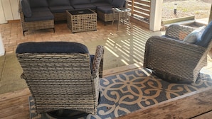 Een overdekt terras met een donkerbank, rieten stoelen en een houten tafel.