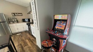 Een keuken met een magnetron, kastjes en een bord waarop 'Welkom' staat. Er staat een basketbalarcadekast en een kruk in de hoek.