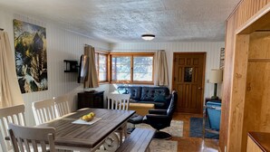Ein Essbereich mit Holztisch, Stühlen und einer Bank. Im Hintergrund befindet sich eine Couch und ein kleiner Tisch mit einer Lampe.
