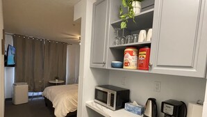 A hotel room with a bed, a small table with a chair, a microwave, a coffee maker, and a shelf with kitchen items.