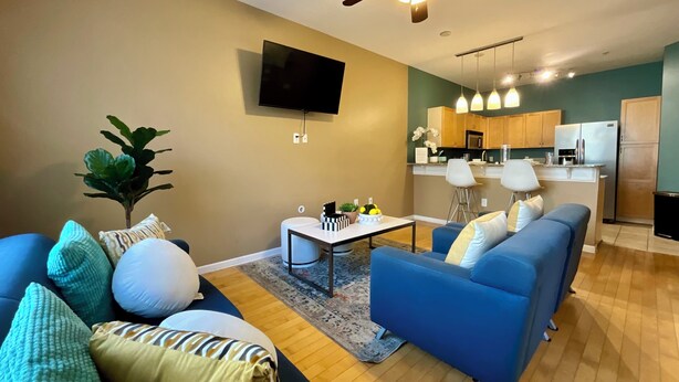Ein Wohnzimmer mit einer Couch, einem Couchtisch und einem Fernseher. Im Hintergrund ist eine Küche mit Kühlschrank und Mikrowelle erkennbar.