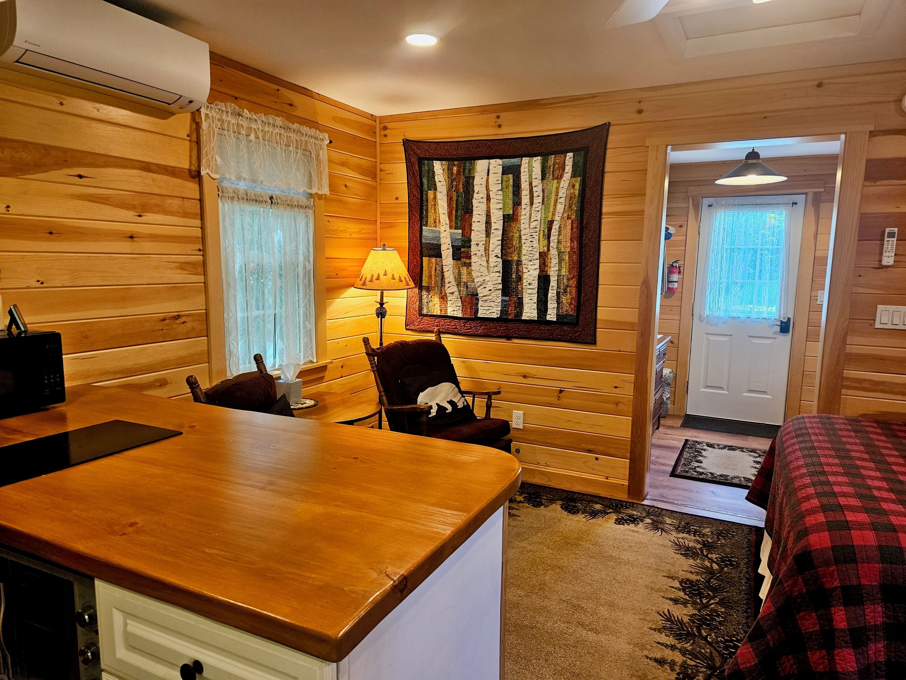 A wooden interior with a dining table, chairs, a lamp, a quilt, and a door.