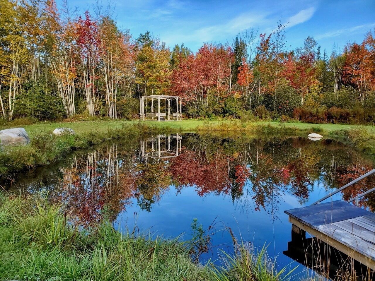 A serene pond with a wooden gazebo and dock, surrounded by autumn-colored trees.