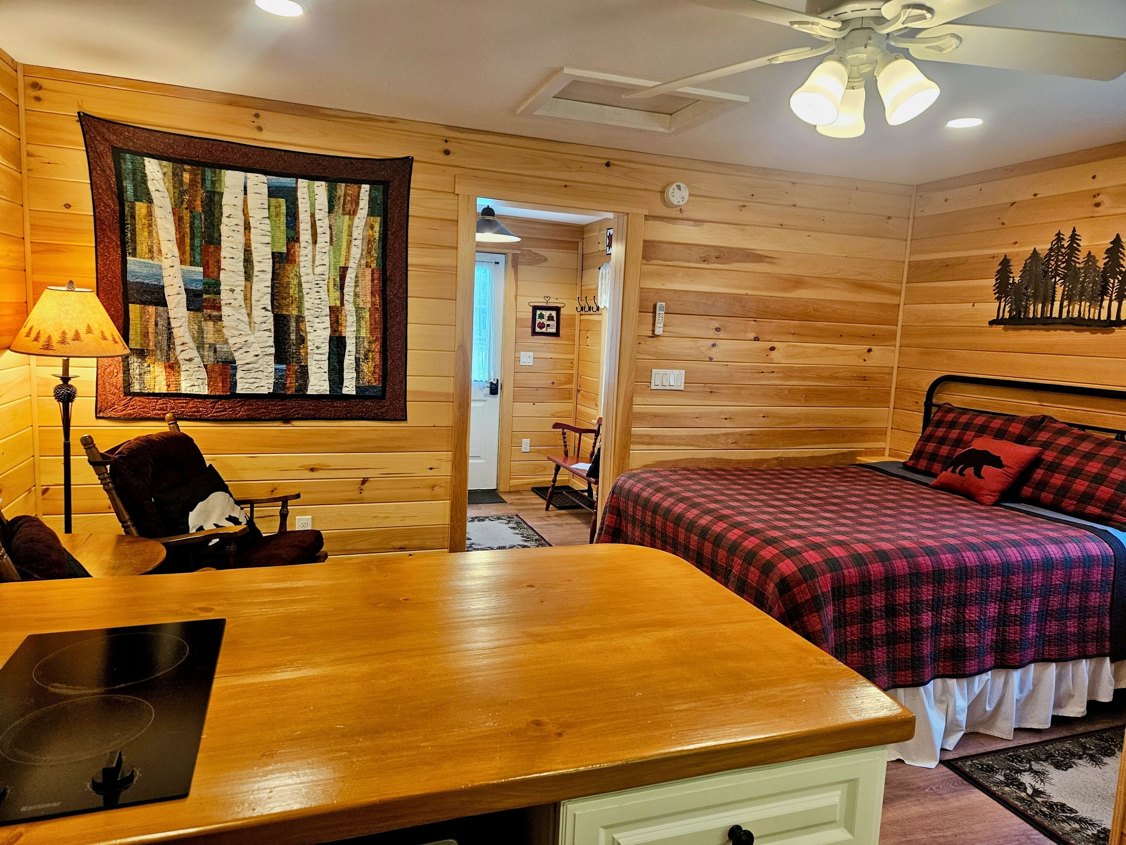 A cozy room with a wooden interior, a bed with a plaid bedspread, a table with a placemat, a chair, and a wall-mounted quilt.