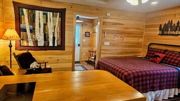 A cozy room with a wooden interior, a bed with a plaid bedspread, a table with a placemat, a chair, and a wall-mounted quilt.