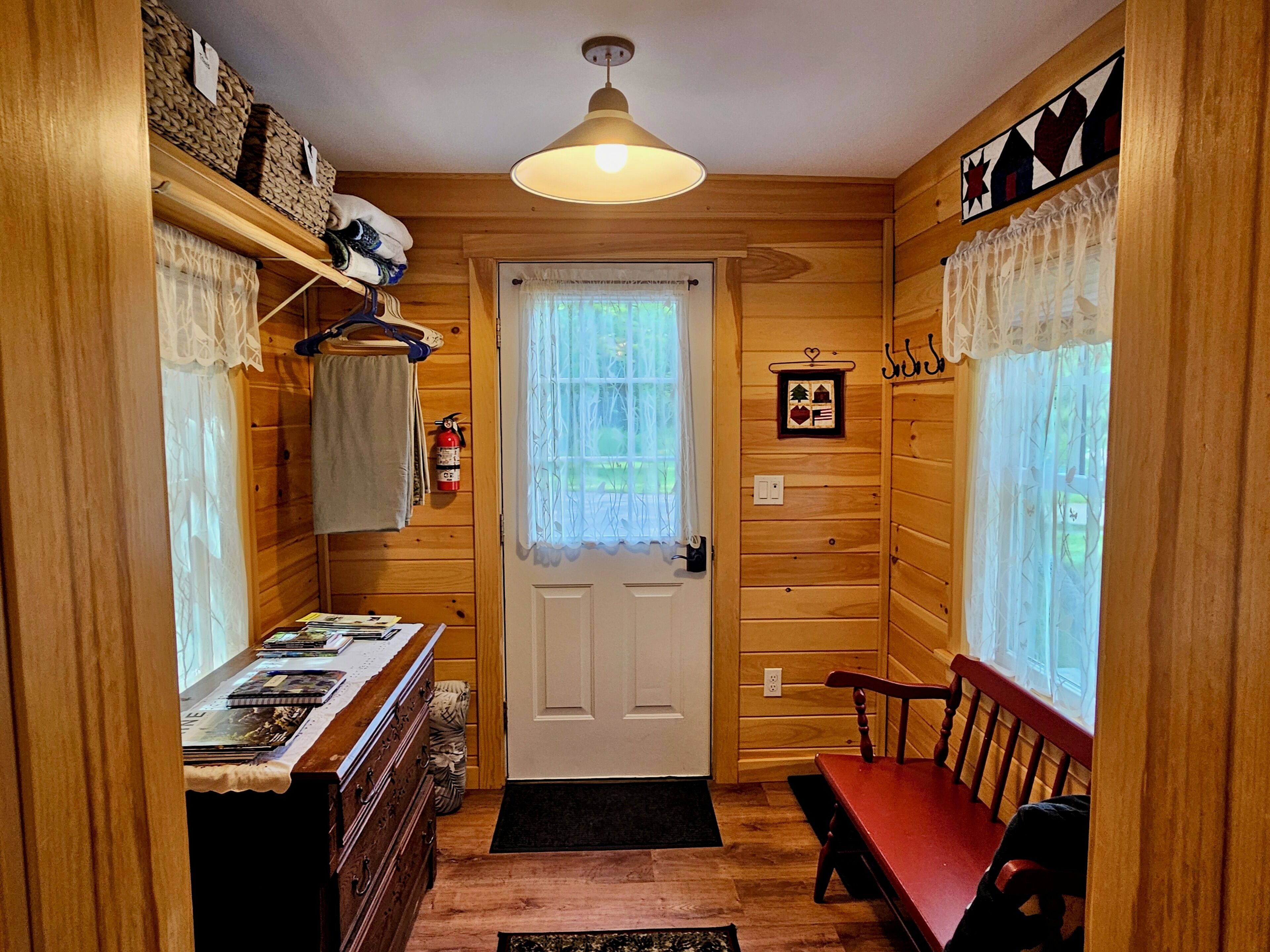 A wooden cabin interior with a door, a table with books, a bench, and a hanging light fixture.