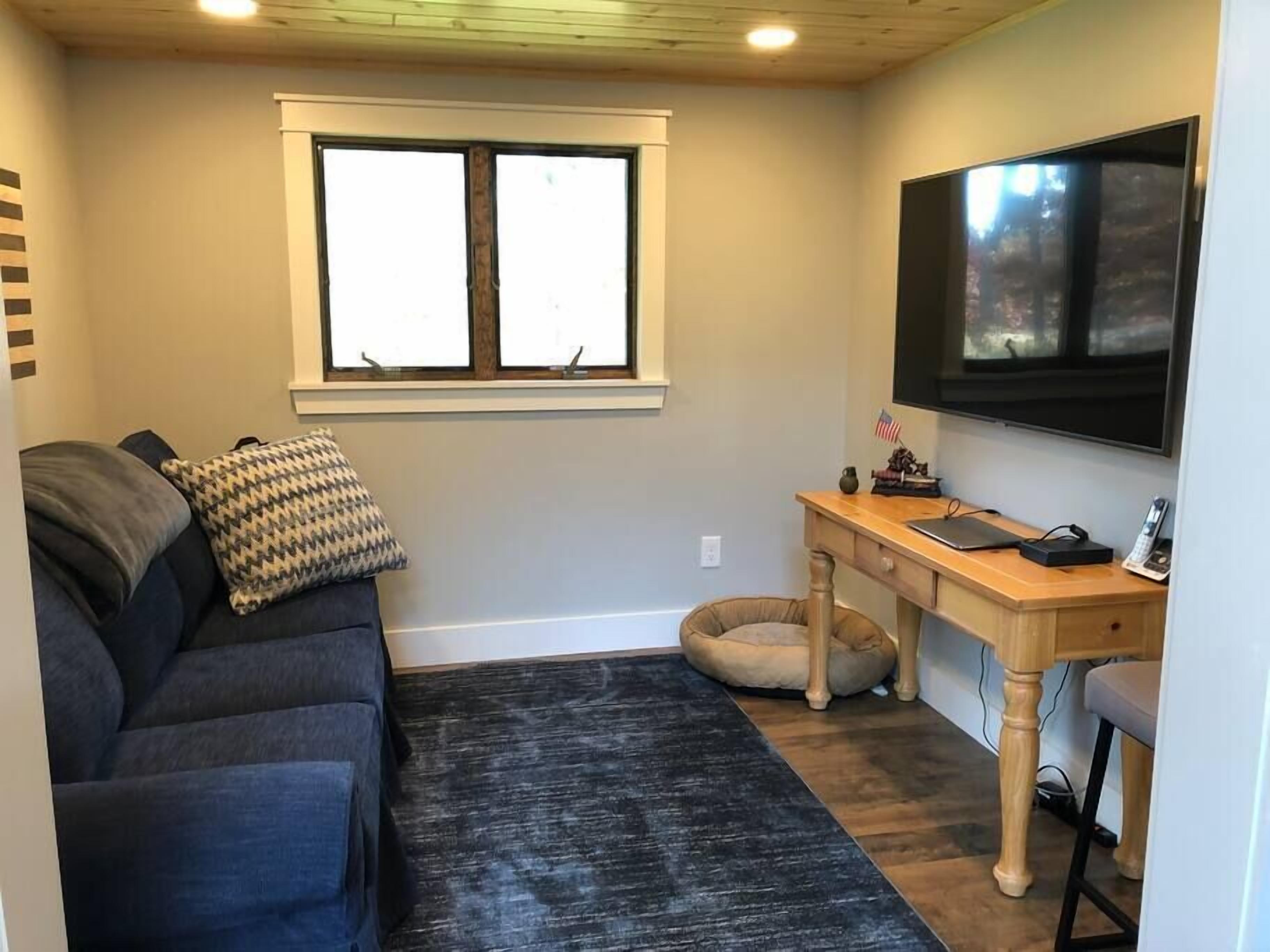 Ein kompakter Wohnraum mit einem dunkelgrauen Sofa, einem kleinen Holztisch, einem an der Wand befestigten Fernseher und einem Fenster über dem Sofa.