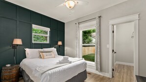 Une chambre avec un grand lit, deux lampes de chevet, une fenêtre avec des rideaux, un tonneau en bois et une porte donnant sur une autre pièce.