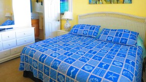 Een slaapkamer met een bed, een kaptafel, een nachtkastje, een lamp en een schilderij van palmbomen.