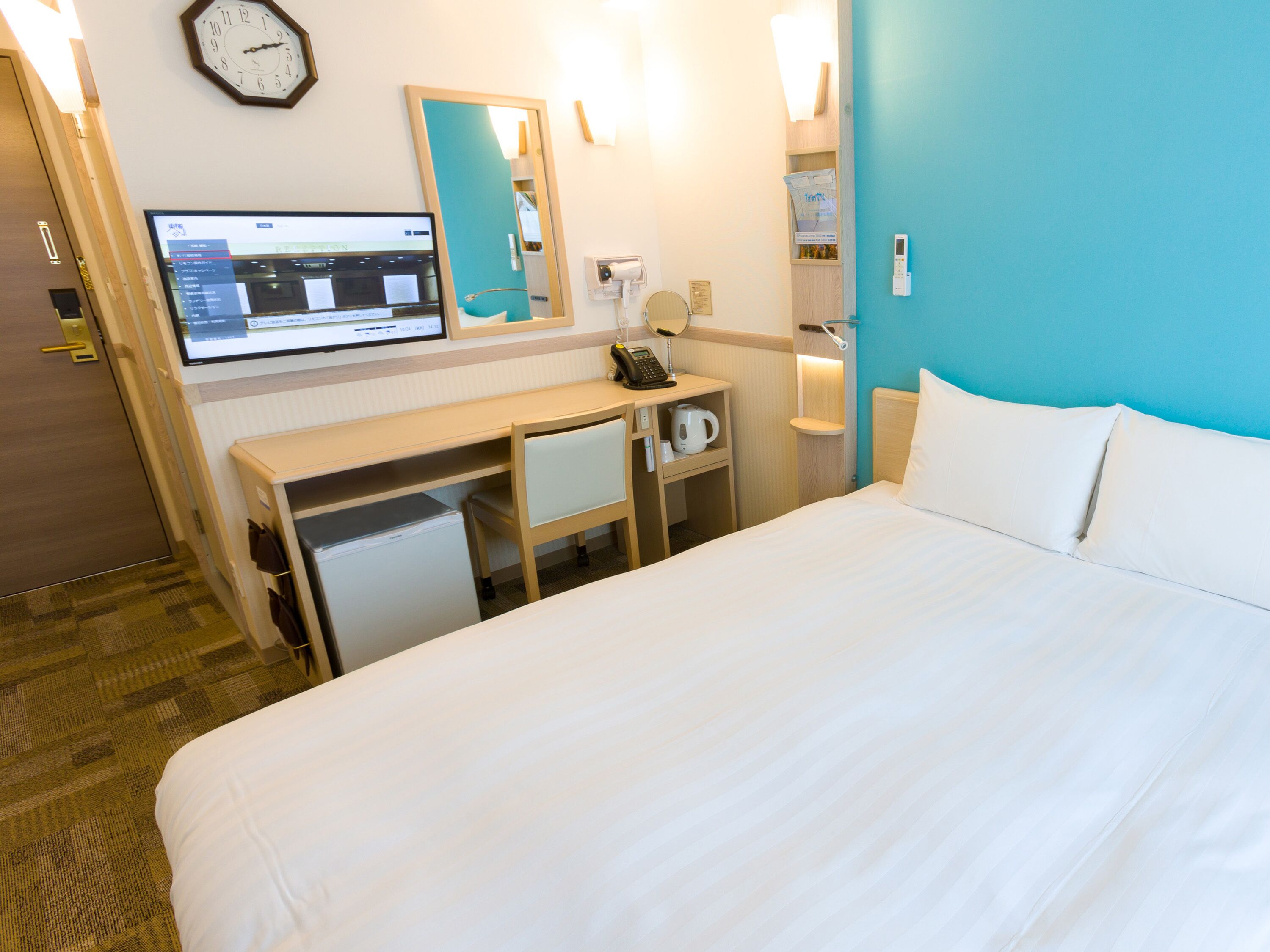 A hotel room with a bed, a desk, a computer, a clock, and a mirror.