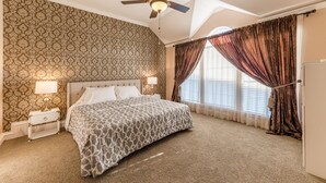 A bedroom with a patterned wallpaper, a ceiling fan, a bed with a patterned bedspread, two bedside lamps, and a window with curtains.