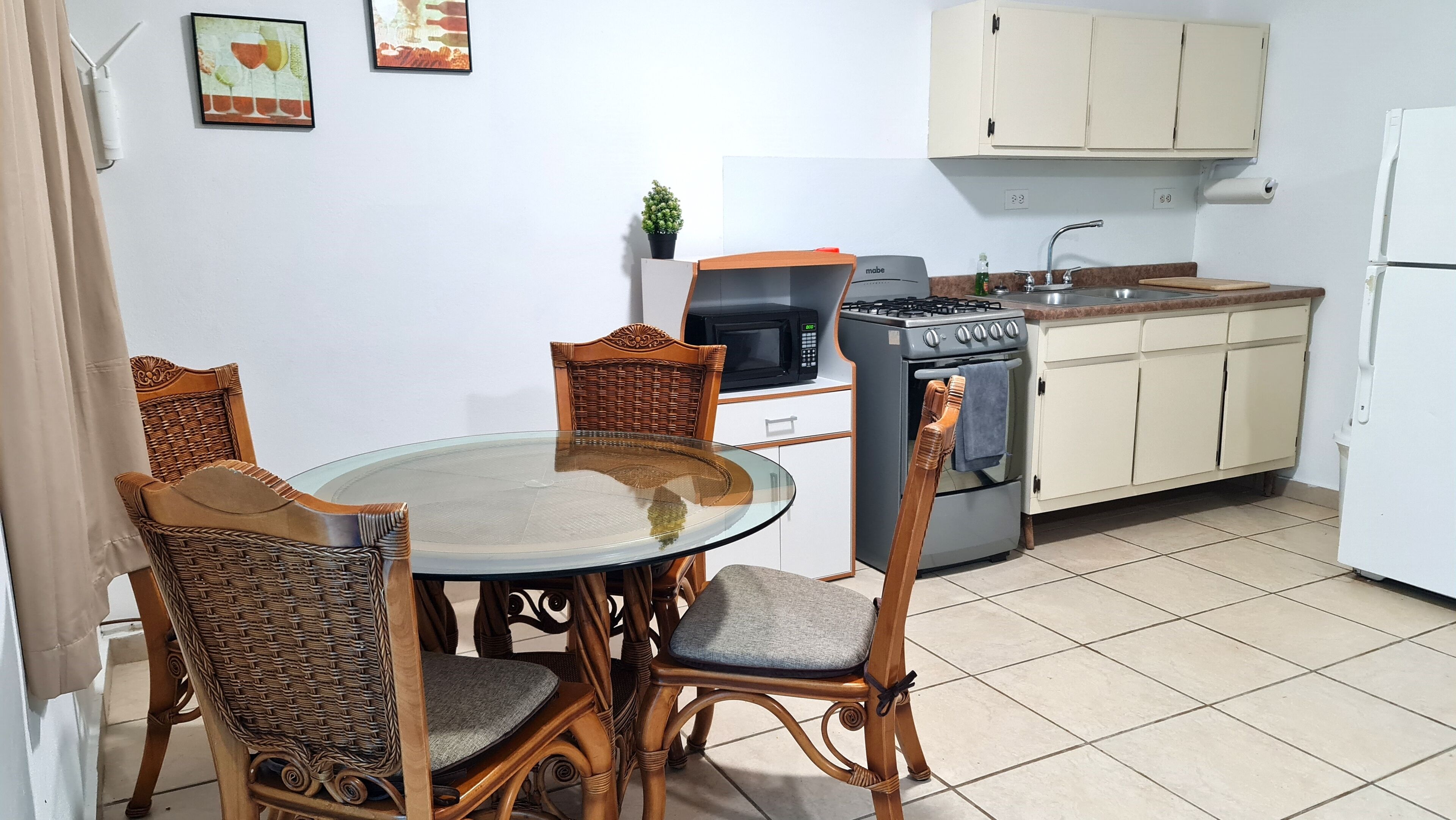 A compact kitchen with a round glass table, wicker chairs, a stove, a microwave, and a small potted plant.