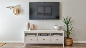 En moderne stue med et fladskærms-tv på væggen, et hvidt konsolbord med bøger og pyngeting, en vævet kurv med en plante, og en vægkunst med hatte.