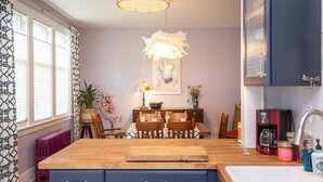 Een keuken met een houten kookeiland, een eettafel met stoelen, een hanglamp en een koffieapparaat.