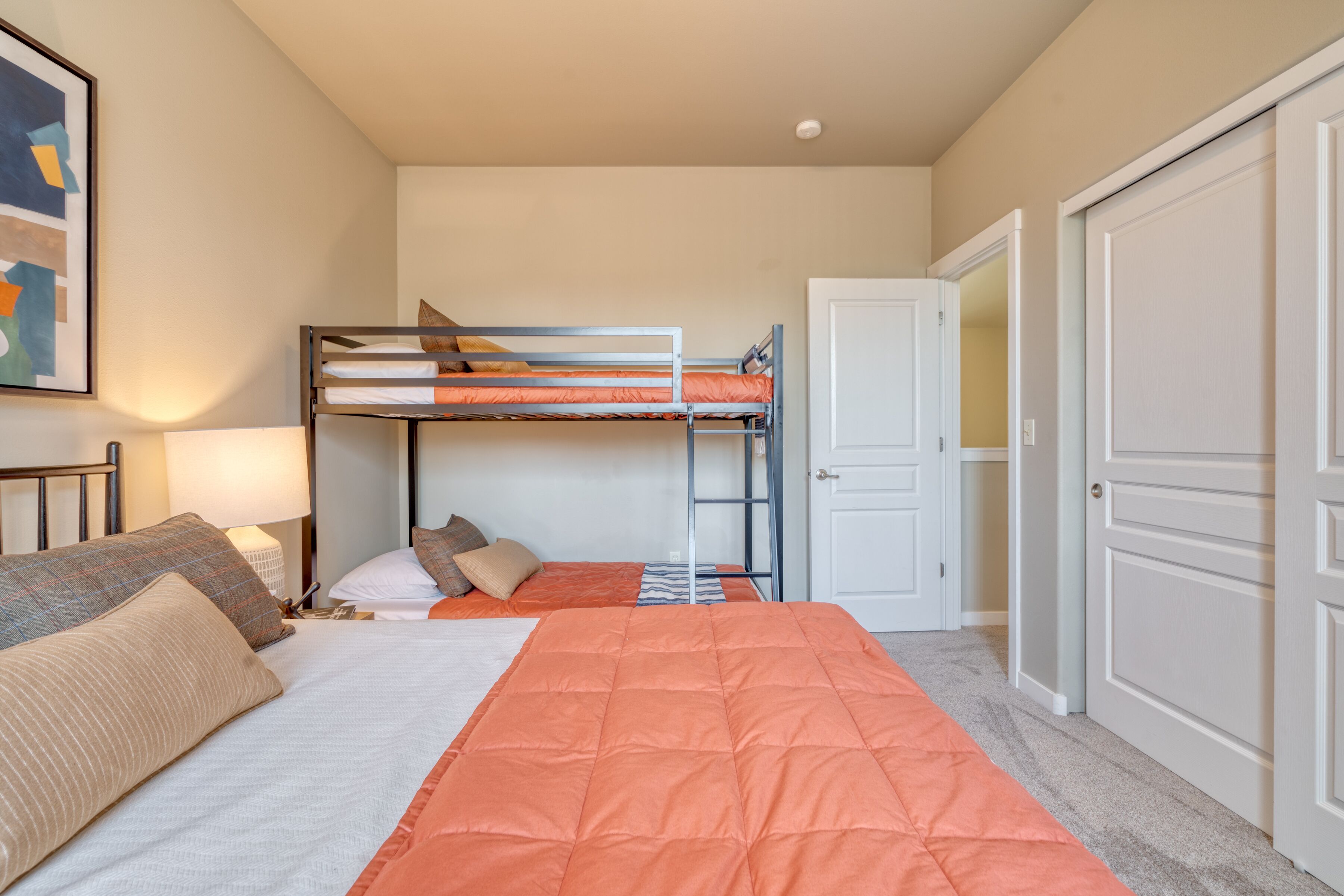 A bedroom with a bunk bed, a single bed, a bedside table with a lamp, and a door to a closet.