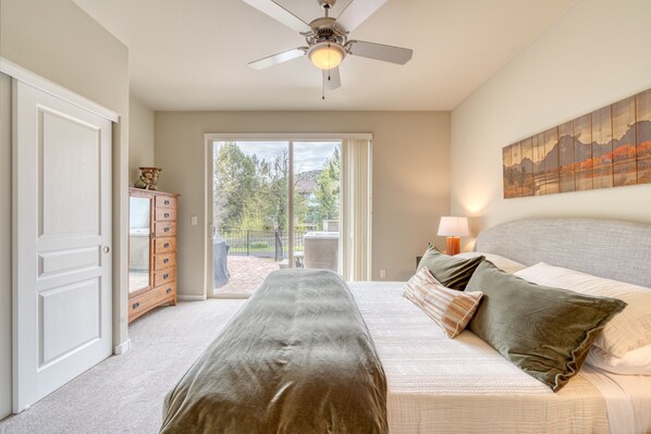 A bedroom with a bed, a ceiling fan, a painting, and a view through a sliding glass door.