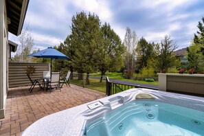 Un jacuzzi en un patio con piso de ladrillo, rodeado de árboles y una sombrilla azul.