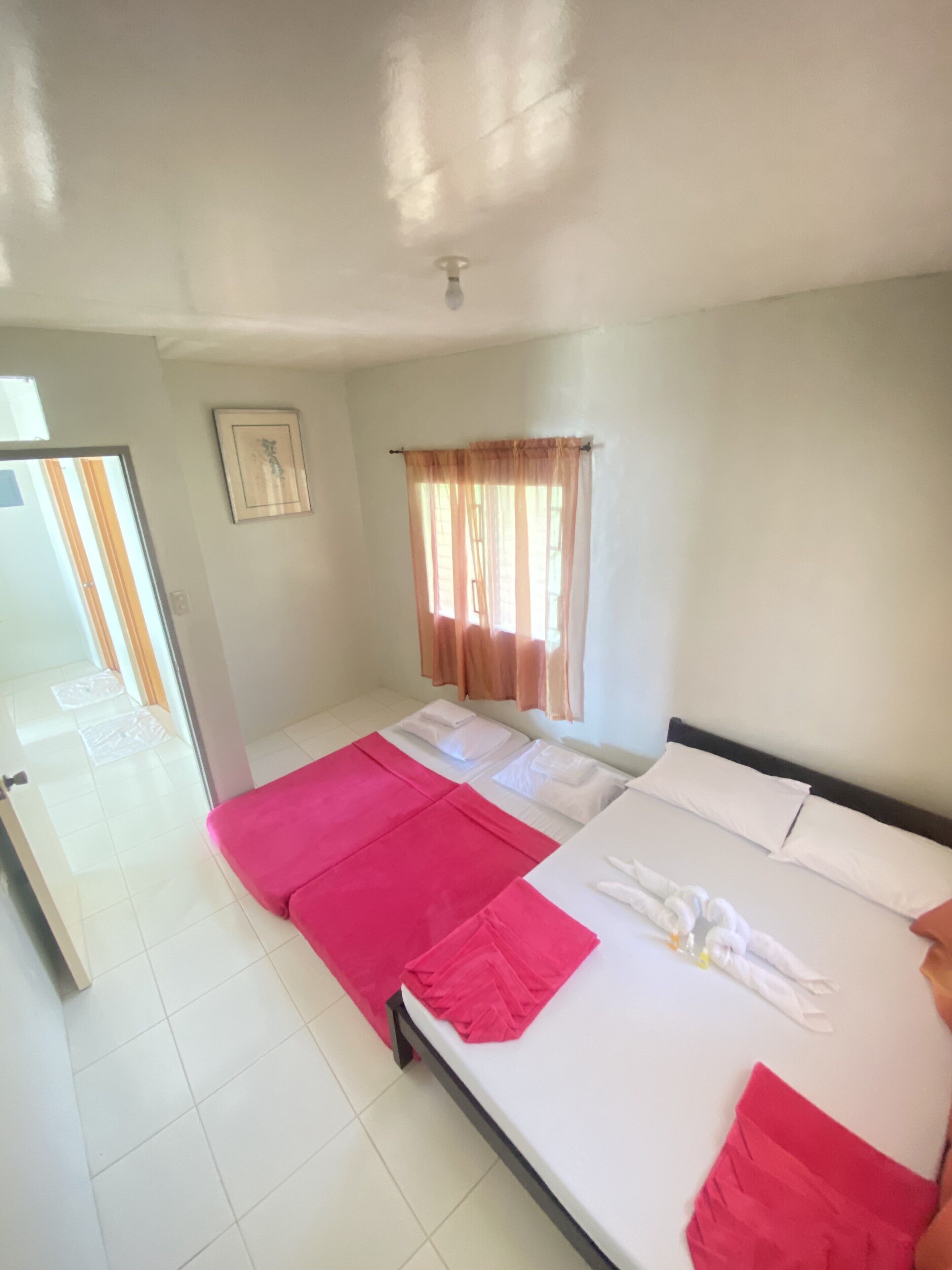 A hotel room with a bed, a window with curtains, a door, and a small picture on the wall.